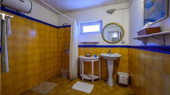 A white & yellow themed bathroom with water heater on the left and shower space beside - Ramgarh Bungalows, Nainital.